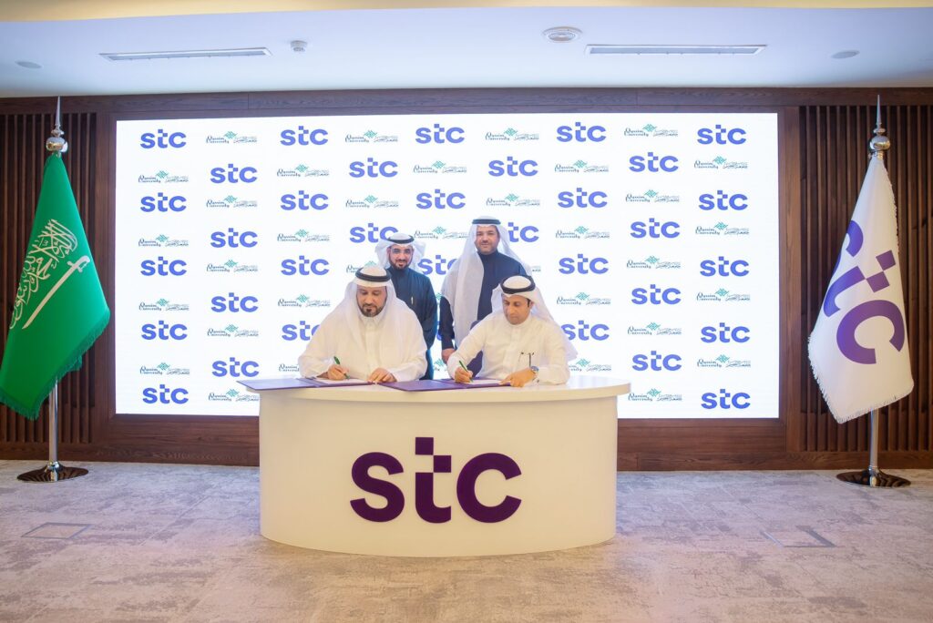 يجتمع أربعة رجال الزي السعودي الأصلي من المجموعة وشماغ أو غترة سعودية لتوقيع تفاهم على طاولة أمام مجموعة شعار STC. أظهر علمان للمملكة العربية السعودية، بينما تتزين الخلفية بعلامات وهي مجموعة مدنية STC، في احترام المواطنة الوطنية للفعالية المنعقدة.
