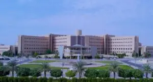 مبنى حكومي فخم وحديث، يتألف من عدة طوابق، ينتصب بشموخ وسط مساحات خضراء منسقة بعناية فائقة وصفوف من أشجار النخيل. يتميز المبنى بمدخل مركزي واسع، يزينه دوار ونافورة أنيقة، تعكس قيم وطموحات رؤية السعودية 2030. يتجلى المشهد تحت سماء زرقاء صافية، حيث يرتدي الزوار وأعضاء هيئة التدريس الزي السعودي التقليدي، بما في ذلك الشماغ أو الغترة، تأكيدًا على التزام المؤسسة بالهوية الوطنية والتميز، بما يليق بمكانة جامعة القصيم.