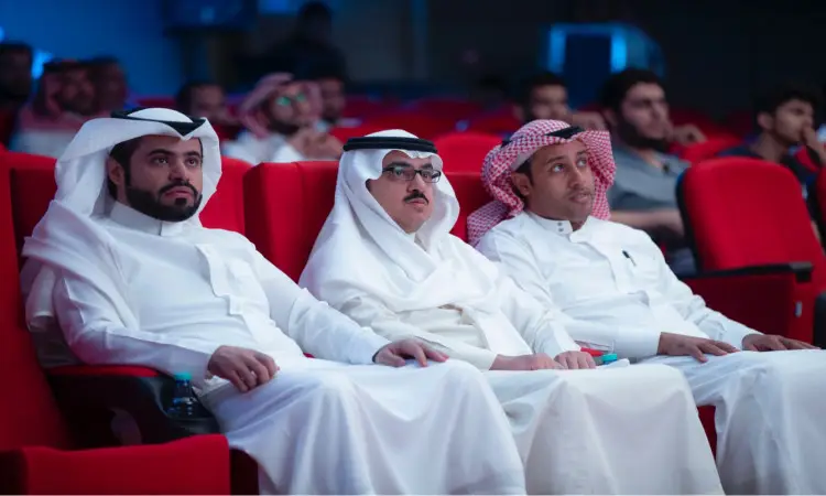 ضبط ثلاثة رجال يرتدون الغترة أو الشماغ والعقال مع البشت السعودي على الصدر الأحمر في المسرح، يتابعون باهتمام ندوة الامتياز. وأشار في الخلفية إلى عدد من الحضور الآخرين، ومن بينهم طلاب العملاق من كلية الصيدلة، وكلهم يبدون اهتمامًا وتركيزًا على ما يُجمَع على مسرح تايلاند.
