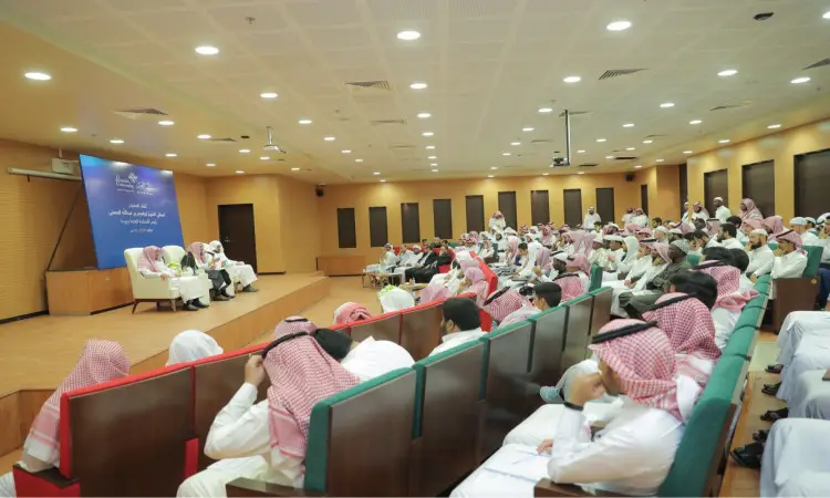 ويظهر في القاعة عدد كبير من الحضور، معظمهم من الرجال الذين تأخروا الغترة أو الشماغ والعقال، بالإضافة إلى البشت السعودي، وهم يعتصمون للاستماع إلى كلمة مدير جامعة القصيم يتقن التكنولوجيا على المنصة، وذلك أمام خلفية زرقاء تتزين بنصوص عربية.