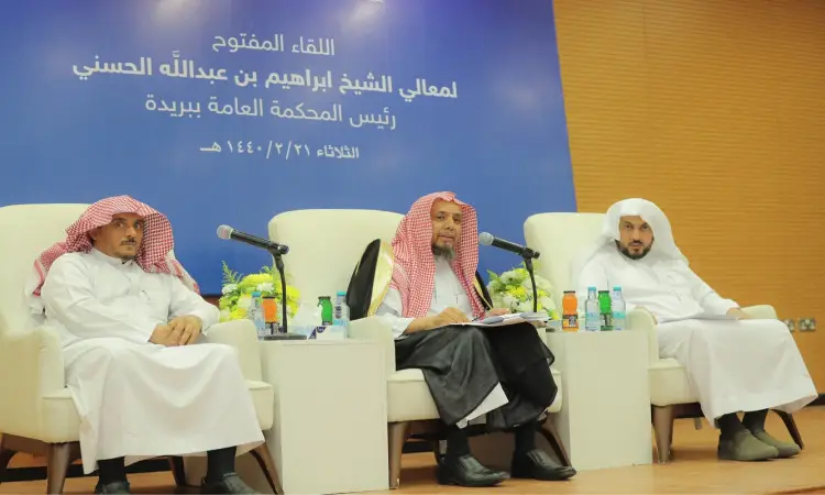 يجلس ثلاثة رجال مرتدين الغترة أو الشماغ والعقال والبشت السعودي على المنصة، وأمامهم ميسات وأوراق وقوارير ماء. وخلفهم على الحائط المكسو بألواح خشبية ونباتية زرقاء تحمل نصًا باللغة العربية يذكر فيها مدير الجامعة. كما تزين الجداول أمامهم باقات من الزهور.