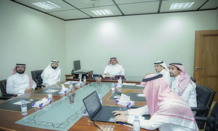 ستة رجال تولى الغترة الشماغ والعقال والبشت السعودي يجلسون حول طاولة ممثلي في مكتب جديد، يبحثن في نقاش حول الدراسات العليا. أمامهم أجهزة حاسب مفتوحة على نظام البلاك بورد، وأوراق موزعة على الطاولة، حيث يشاركون جميعًا بشكل تعاوني في الاجتماع الرسمي ضمن جامعة القصيم.