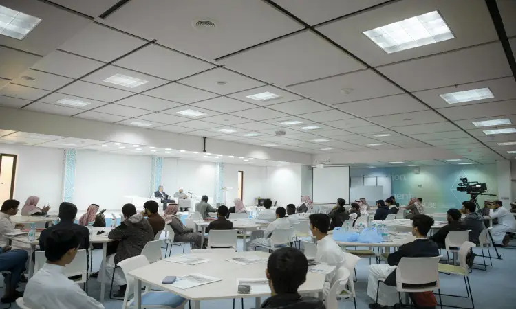 يجتمع مجموعة من الأشخاص، معظمهم من الرجال الذين يرتدون الغترة أو الشماغ والعقال مع البشت السعودي، حول اجتماعات متجولة في قاعة المؤتمرات الحديثة والمضيئة بكلية الاقتصاد والإدارة في جامعة القصيم. ويستمع بالحضور باهتمام إلى القاعة الأساسية، فيما تقوم الكاميرا بتوثيق مجريات الفعالية.