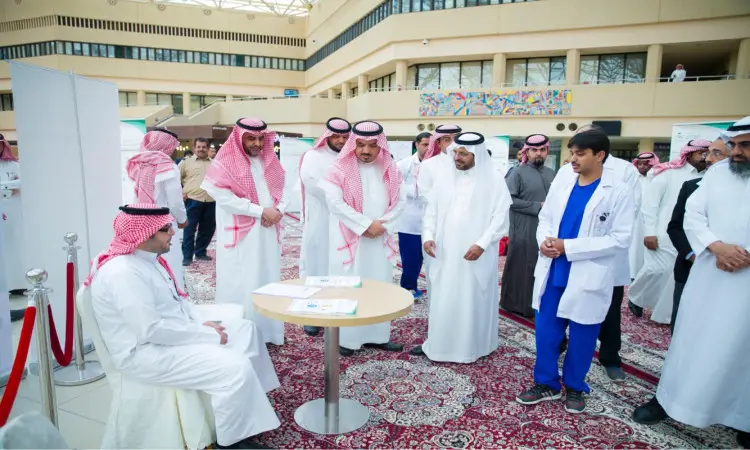 تجمّع عدد من الرجال، منهم مدير الجامعة، في مقر المدينة الجامعية أثناء حملة التطعيم ضد الإنفلونزا الموسمية. وقد ارتدى بعض الحضور الغتر السعودية أو الشمغ مع العقال والبشت السعودي، بينما كان كان بزيهم الطبي الرسمي. وأجرى التفاعل بين المشاركين حول طاولة مواضيعية تعريفية وتوعوية صعبة بالحملة.