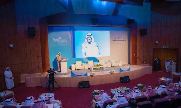 رجل يرتدي البشتاء السعودي والغترة والعقال على المنصة في القاعة حديث تتوزع فيه الرجال وتزينه الزهور، فتحة بكلمة البحث العلمي في آسيا، بينما يستمع يستمع بلباسهم الرسمي من شماغ سعودي وعقال في أرجوة. تسجيل صورته الحديثة على شاشة كبيرة في مختبر القصيم في المملكة العربية السعودية.