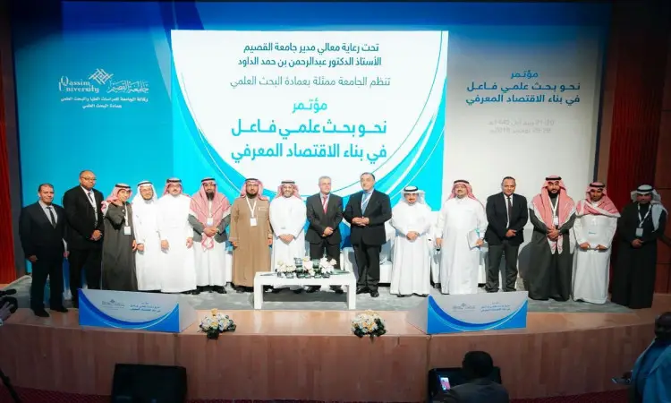 القطة مجموعة من رجال مرتدين الغترة أو الشماغ والعقال والبشتت السعودي، إلى جانب سيدات بزيليغ، على منصة المسرح، صورة جماعية خلال أحد المؤتمرات العلمية في جامعة القصيم. يبرز أهمية البحث العلمي في تطوير اقتصاد الصناعة ومستلزماته بالعربية.
