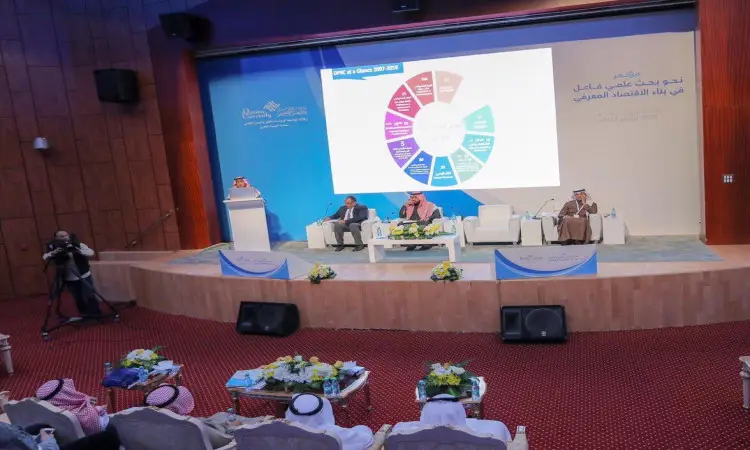 أعضاء لجنة المؤتمر في المسرح، مرتدين الغترة أو الشماغ السعودي مع العقال، بالإضافة إلى البشت السعودي الرسمي، أمام شاشة كبيرة تعرض بشكل دائري ملوناً. يناقش الأعضاء منشوراً بشكل علني هاماً في الحضور، بينما يُنصت الحضور باهتمام داخل قاعة مزينة بالألواح الخشبية وأرضيات مفروشة بسجاد أحمر، وذلك في رحاب جامعة القصيم.