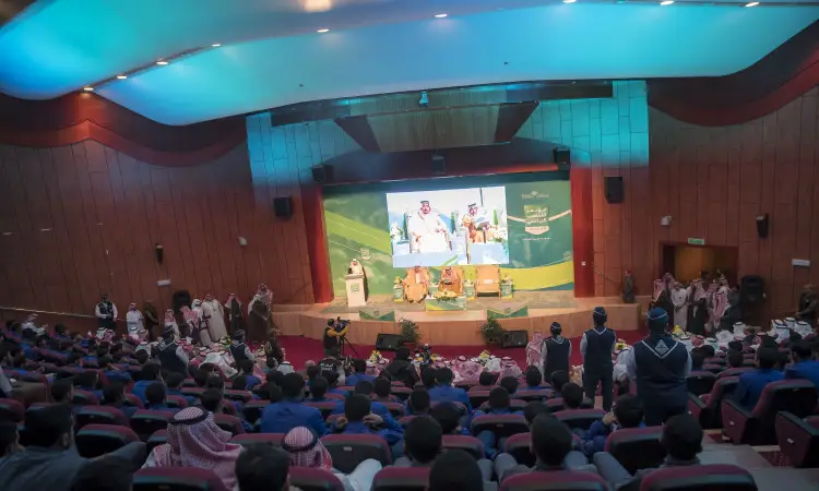 يجتمع جمع كبير من الحاضرين في قاعة محاضرات في القصيم، حيث يتابعون عرضًا تقديميًا على التيس حول موضوع "التعصب الرياضي: الآثار والحلول"، يقدمه عدد من التعريفين من منصة الخطابة. ويتم عرض على الشاشة صور للأشخاص زيًا تقليديًا تشمل الغترة السعودية أو الشماغ والعقال، بالإضافة إلى البشت السعودي. ويلاحظ أن كثيرًا من تأخرين الزي الرسمي للجامعة أو اللباس التجريبي السعودي تفاصيله، مما يضفي طابع الوقار والجدية على الفعالية العلمية المنعقدة في رحاب الجامعة.