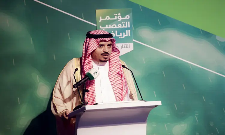 ارتدى الرجل الغترة والعقال والبشتاء السعودي غالبا من على المنصة خلال مؤتمر التعصب الرياضي، وذلك في القصيم. ساهم في الخلفية نص عربي وتصميم أخضر. وتوجد على الموقد شماعتان للميكروفون المخصص لمعالجة الكلمة.