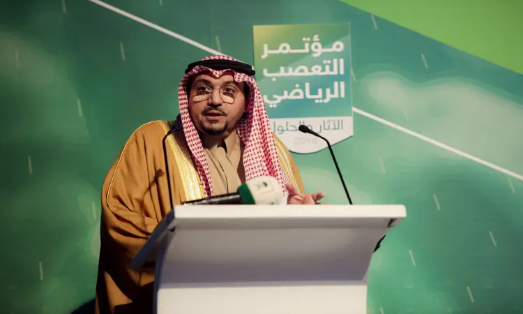رجل يرتدي الغترة والشماغ والعقال والبشت الرسمي السعودي، مشهورًا في مؤتمر المنصة في القصيم. ضع خلفه لوحاً أخضر بالضغط على النصوص باللغة العربية، بينما يوجد أمامه ميكروفون يُبرز كلمة "أمير القصيم" والجهود التي تتناول الحساسيات الرياضية.