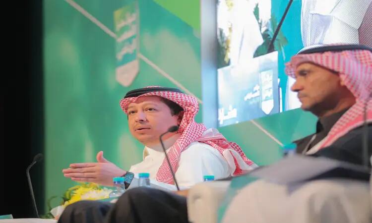 يجلس رجلان يرتديان الغترة السعودية مع العقال، وأحدهما يرتدي بشتًا سعوديًا فوق ثوبه، على منصة النقاش ضمن مؤتمرات مؤتمر التعصب في القصيم. بوضوح لا أحد يقصد مشيرًا بيده أثناء مناقشة دور الإعلام في إثارة التعصب، بينما يستمع الآخر باهتمام. تسليط الضوء على خلفية المسرح الخشبية أمام خلفية خضراء، تعلوها شاشة كبيرة تعرض صورهم مباشرة للحضور.