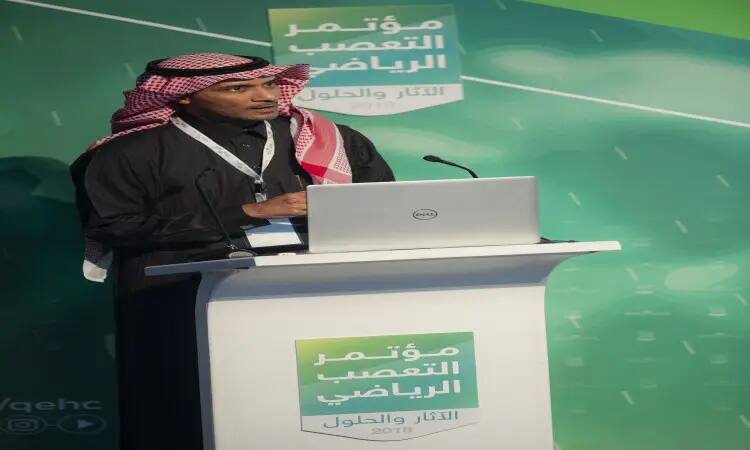 رجل القط مرتدياً الغترة والعقال والبشت السعودي خلف منصة لابتوب، يلقي كلمة. استماع على المنصة وفي خلفية كتابات باللغة العربية متحد بمؤتمر التعصب لعام 2018 دون الإعلام في تمام للتعصب الرياضي، وذلك ضمن فعاليات جامعة القصيم.