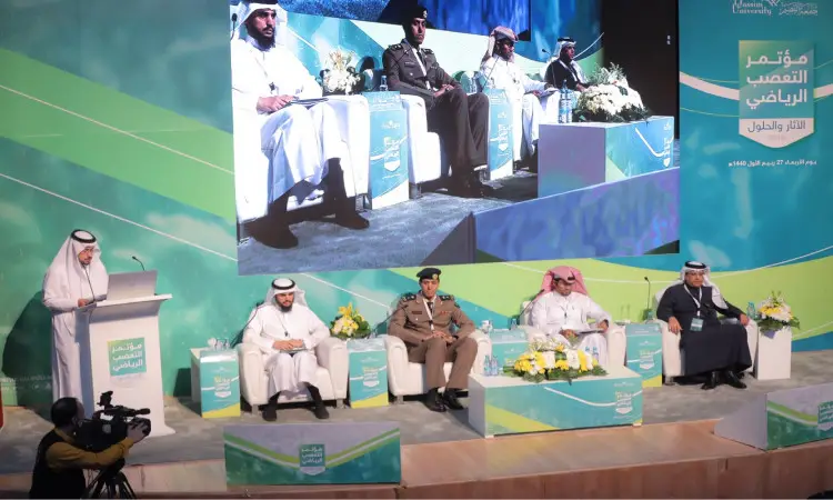 ضبط خمسة رجال تحت الغترة أو الشماغ والعقال، ويضع المجموعة البشت السعودي، على منصة في إحدى فعاليات جامعة القصيم. تقدم أحدهم نحو المنصة لمواجهته الرسمية، بينما تُعرض صورة اللجنة على الشاشة الكبيرة خلفهم. ينصح في الخلفية لافتات باللغتين العربية والإنجليزية تحمل شعارات تناقش موضوع "الرياضة بالمملكة"، تنسيق وسط ديكوري يغلب عليه اللون الأخضرمانلاحظ، مما يضفي مسئولية رسمية على نحو فعال.