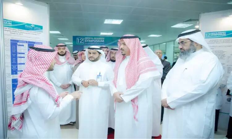 مجموعة من الرجال الحاضرين الغترة والشماخ والعقال، بالإضافة إلى البشت السعودي التقليدي، يتبادلون النقاش حول ملصقات بحثية خلال فعاليات الملتقى الطبي البحثي في القصيم، للشرب في لوحات إرشادية تشير إلى الخلفية "الملتقى الطبي البحثي الثاني عشر" و"مدير الجامعة".