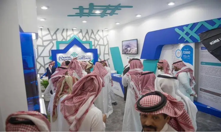 A group of men resplendent in Saudi bisht, vest, and shamma gathered inside a modern pavilion at the 33rd Janadriyah Fair, whose walls are adorned with a blue geometric pattern, as well as informational panels about Qassim University and its affiliated veterinary clinic.