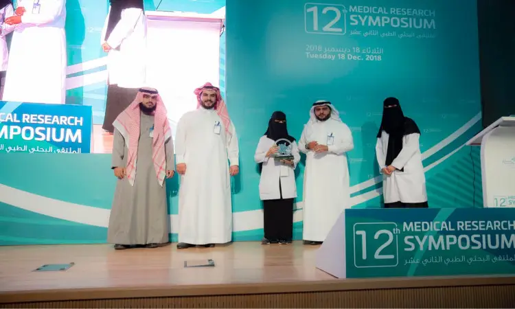 لست خمسة أشخاص مرتدين الغترة أو الشماغ والعقال والبشت السعودي على منصة الملتقى البحثي الطبي الـ12 في جامعة القصيم. وتسلّم إحدى الجوائز العلمية عن الثالث، بينما يتابع الحضور من اللجنة العلمية ذلك، لإعداد خلفهم تفاصيل الأحداث وشعار اللجنة على العرض.
