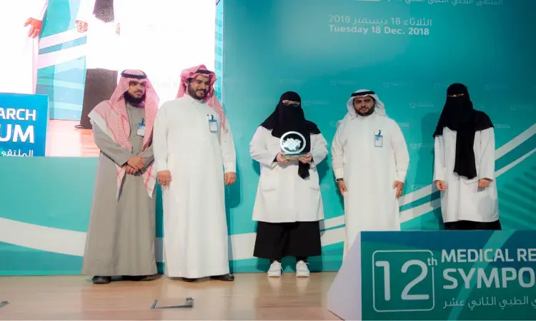 ستة أشخاص يرتدون الزي السعودي الرسمي على منصة ملتقى البحث الطبي في القصيم؛ حيث يظهر الرجال وهم وهم الغترة أو الشماغ مع العقال والبشت السعودي. فازت بجائزة الفائزة في المجال الجديد وهي تحمل جائزة التكريم. تسجيل تفاصيل الفعالية وعبارة "الملتقى الثاني عشر للبحوث الطبية" على لافتات زرقاء في خلفية المنصة.