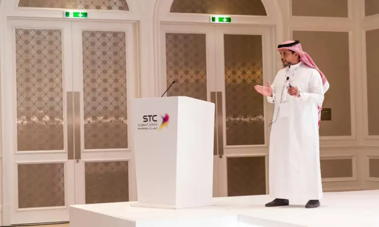 رجل الاتصال مرتدياً غترة سعودية وعقالاً وبشتاً سعودياً على المنصة بجانب منبر عصر شعار STC، مقدماً عرضاً رسمياً حول اشتراك مع STC، وذلك في قاعة مؤتمرات ذات أبواب ومزدوجة أنيقة.