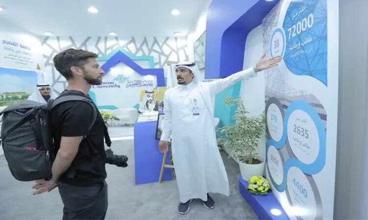 في جناح جامعة القصيم بمعرض الجنادرية 33، يرتدي رجل يرتدي غترة سعودية وعقال وبشتلي وهو يشير بيده نحو شاشة عرض معلوماتية، تومكسًا إلى زائر يرتدي لباسًا عمليًا ويحمل الكاميرا. ويعرض الجناح التنفيذي للجامعة وإنجازاتها في الوقت الذي يملؤها الحضور، وشارة التعريفية والترويجية للجامعة.