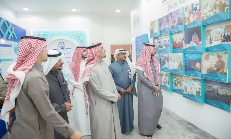 الاتصال عدد من الرجال بلباسهم التقليدي، مرتدين الغترة أو الشماغ السعودي مع العقال والبشت السعودي، في صالة عرض خلال فعاليات الجنادرية 33. يستخدمون باهتمام جداراً مخصصاً لاستهداف الصور واللوحات التعريفية التي تُبرز عملات وهمية جامعة القصيم وإنجازاتها المتميزة عبر السنين.