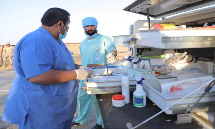 Two health care subby, dressed in medical uniforms with a Saudi shemagh, sheqal and besht, prepare medical supplies on a mobile cart in the open air near the camel fields during the 33rd Janadriyah events, and contribute a number of medical devices and bottles to the cart. Behind them can be seen the desert and some participants from the Qassim University Veterinary Clinic.