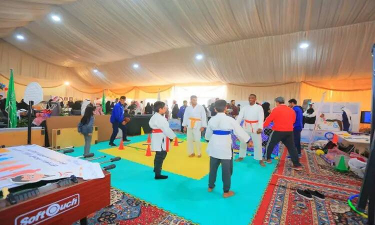 يتدرب مجموعة من الأشخاص، بما فيهم الأطفال مرتدون زي الكاراتيه الرسمية، على الفنون القتالية فوق بساط مخصص وسط حيث مفعمة بالحيوية والألوان في مخيم الجامعة خلال فعاليات ربيع بريدة. الخلفية، يتجوّل الزوار الذين يحضرون الغترة أو الشماغ مع العقال والبشت السعودي بين الأجنحة وأيكان المعدات الرياضية، مستمتعين بالتجربة الثرية وفي تقديم جامعة القصيم لمجتمعها وزوراها الكرام.