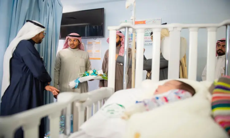 A number of men wearing a Saudi ghutra or shemagh with an aqal, as well as a Saudi bisht, near the hospital, chatting about medicine. Squeezing one of the flowers, while a child lies in bed, partially covered by a blanket.