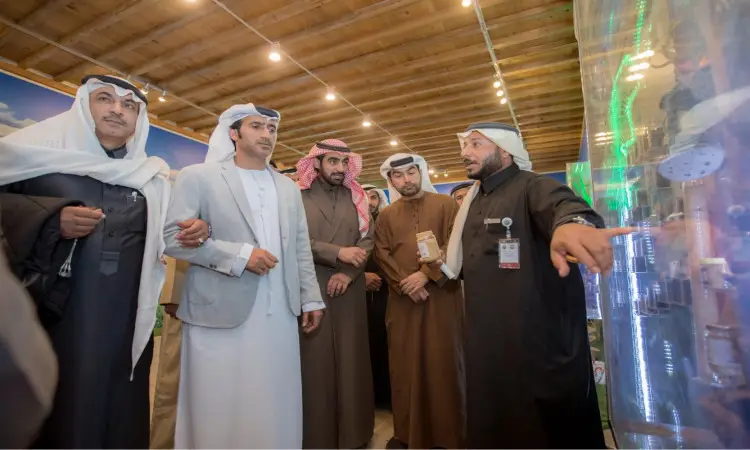 مجموعة من الرجال تولى الغترة أو الشماغ والعقال والبشت السعودي يجتمعون للاستماع إلى رجل، ويرتدي كذلك الزي التقليدي السعودي، وهو يشير إلى عرض عناصر في قاعة التصميم الحديثة ضمن فعاليات مهرجان الجنادرية، وذلك تحت سقف الخشب.