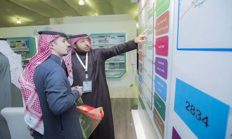 يتفحص رجلان يرتديان الغترة السعودية والعقال والبشت السعودي لوحة معلومات في معرض القصيم للكتاب؛ حيث يشير إلى تفاصيل حول محتويات مكتبة واضحة عبر مخططات ملوّنة ونصوص باللغة العربية.