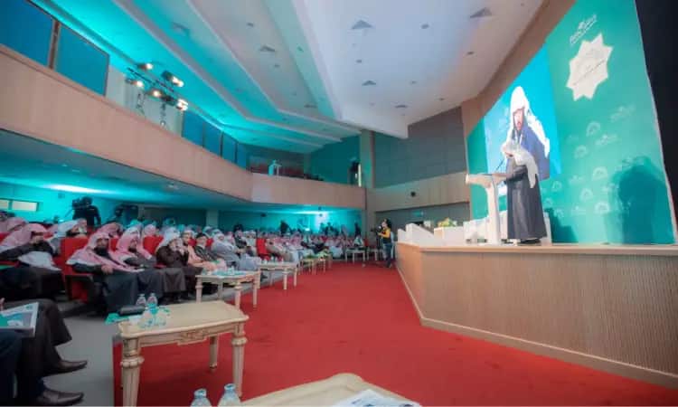 معطف يرتدي الزي السعودي التقليدي، مشتملاً الغترة والعقال والبشت، خلف منصة الخطابة على المسرح في قاعة ذات إضاءة زرقاء ساطعة وسجاد أحمر أنيق. ويخاطب هذا المكان جمعاً من الحضور في جامعة القصيم، يتسمون بالجدية ويتوقع بقضايا البحث العلمي. وقد ارتدى العديد من الحديثين عن الشماغ السعودي أو الغترة السعودية، مغغين للمتحدث باهتمام الكبار وحرص أكاديمي.