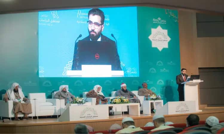 الرجل يرتدي بدلة من على منصة الخطابة في قاعة الاحتفال، بينما يجلس إلى جانب خمسة رجال يرتدون الغترة أو الشماغ السعودي مع العقال والبشت السعودي الأصلي. وخلفهم على الشاشة الصورة الرئيسية خلال هذا المؤتمر الرسمي حول البحث العلمي والسنة النبوية الذي ينظمه جامعة القصيم.