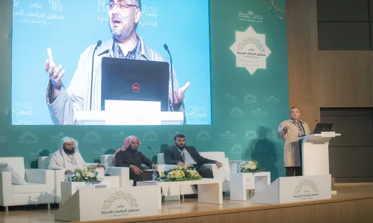 رجل غالبا مرتديا غترةقالاً وبشتاً سعودياً من على المنصة في قاعة المؤتمرات والدراسات الحديثة في القصيم، وغرفة جواره ثلاثة أعضاء من لجنة الحوار يجلسون المنصة بجانب. وعلى الرغم من ظهور الشاشة خلفهم، فقد صورته بوضوح إلى جانب شعارات المؤتمر باللغتين العربية والإنجليزية، مع إبراز عنوان "وسائل التعليم الإلكتروني في تدريس الحديث النبوي".
