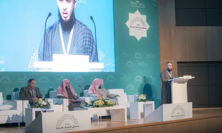 يلقي أحد كلمته على المنصة خلال مؤتمر الدراسات الحديثة في القصيم، مرتدياً الغترة والعقال والبشت السعودي، بينما يستمع إليه ثلاثة من أعضاء اللجنة – جميعهم وتبقى الشماغ السعوديشت – باهتمام. تمت الإشارة إليه على شاشة كبيرة خلف الجدول، ويبرز في الخلفية نصٌ باللغة العربية متعلق بتدريس الحديث النبوي وشعارات المؤتمر، مع التنسيقات من الورود الموزعة على الجداول باستثناء الطابعة الرسمية للمناسبة.
