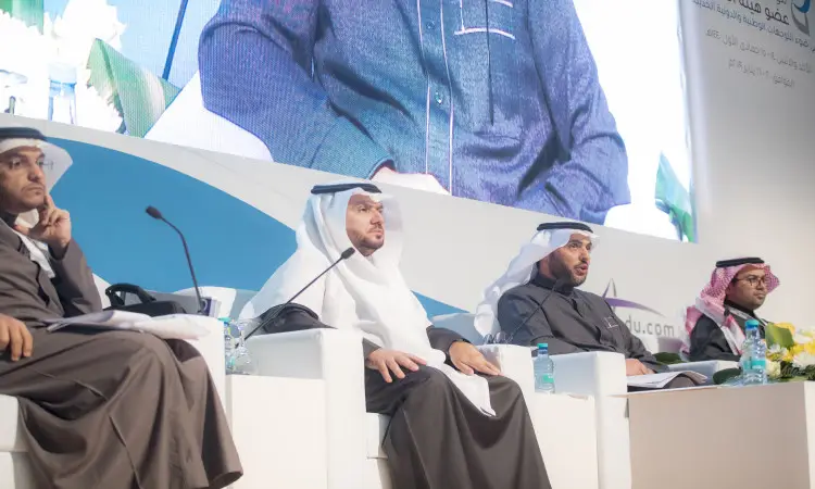 أربعة رجال يرتدون الغترة أو الشماغ والعقال بالإضافة إلى البشت السعودي على منصة نقاش ضمن فعاليات المؤتمر الدولي لتقويم أداء عضو هيئة التدريس في جامعة القصيم، وأمامهم ووثائق وزجاجات مياه. احترس من خلفهم شاشة كبيرة تم التقاط صورة مكبرة لشخصين في الجلسة.