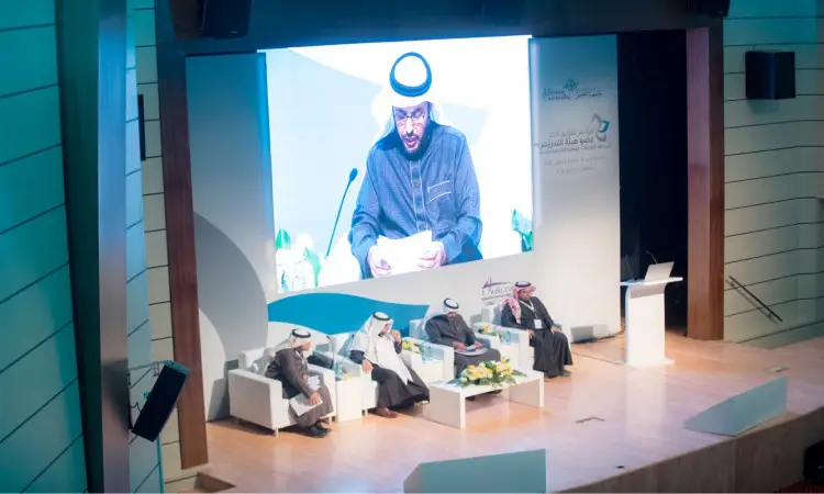 يجلس أربعة رجال غترة أو شماغًا سعوديًا مع عقال وبشت سعودي على المنصة خلال جلسات المؤتمر، بينما يقوم أحد بإلقاء كلمته من على المنبر. يجب أن يكون على الشاشة الكبيرة خلفهم صورة لمتحدث وثائقي من أوراق البيتزا أثناء فعاليات المؤتمر الدولي لتقويم أداء عضو هيئة التدريس في جامعة القصيم.