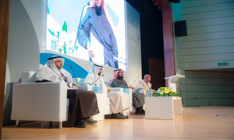 يجلس الرجال الأربعة تحت الغترة السعودية والعقال والبشت السعودي على منصة المؤتمر الدولي في قاعة القصيم، يناقشون تطوير الأستاذ الجامعي، وأمامهم وقوارير. وعرضوا خلفهم شاشة كبيرة تحمل صلة بالموضوع، ما يعكس أنها مؤتمر أو صورة ندوة علمية للأطفال.
