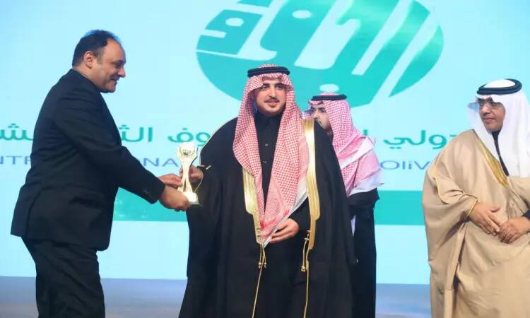 A man in a suit hands an award to a man dressed in a traditional Saudi outfit consisting of a Saudi shemagh and a Saudi bisht, possibly a faculty member, on stage. Alongside them are two other figures and a large Arabic script, as well as a large logo - the backdrop event appears to be part of Creative Intelligence under the leadership of His Highness the Emir of Al-Jouf region, in the halls of Al-Qassim University.