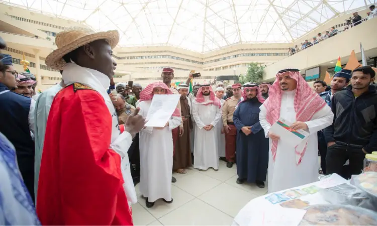يرتدي رجل غترة سعودية وعقالًا وبشتًا أحمر اللون يقوم بقراءة ورقة نص من أمام مجموعة من الحضور، حيث يظهر العديد منهم مرتدين الزي التقليدي وعلى رأسهم الشماغترة والبقالشت السعودي. جاء هذا التجمع في بهو واضح تعلوه قبة، وذلك من خلال مهرجان تراث الشعوب الذي اضافته جامعة القصيم وشاركوا في تمثيل أكثر من دولة.
