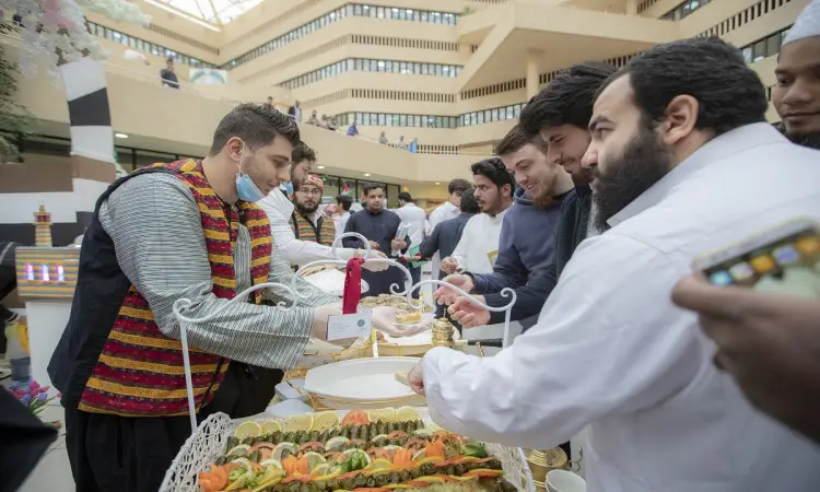 اتصل بمجموعة من الرجال، من بينهم الطلاب، مصطفين عند منصة توزيع الطعام داخل الصندوق. ودعا مقدم الطعام البشت السعودي مع الشماغ والعقال إضافة إلى الكمامة الطبية أثناء تقديم الأكلة. تزين أطباق تقليدية تقليدية وأصناف متنوعة من الخبز، وذلك ضمن فعاليات مهرجان تراث الشعوب في القصيم.