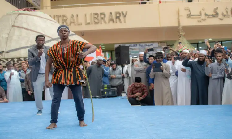 رجل حافي القدمين مرتديًا الأصلي الأفريقي، حامل عصا، على منصة زرقاء. وأمام مكتبة الجامعة المركزية، تومض التلاميذ الصغار وهم الشماغ السعودي أو الغترة مع العقال والبشت السعودي ويوثقون اللحظة بالتصوير احتفاءً بتراث الشعوب في هذا الحدث الذي نظمته جامعة القصيم.