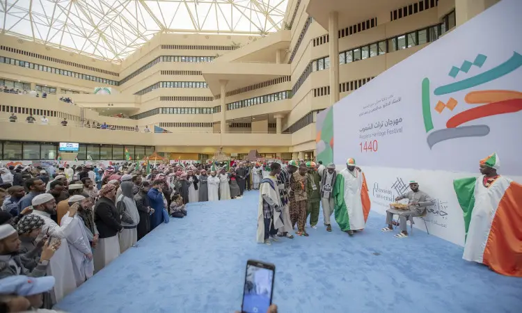 يتجمع عدد كبير من الحاضرين في قاعة حول منصة زرقاء، حيث يشارك طلابنا بوبارتين الغترة أو الشماغ السعودي مع العقال والبشت السعودي في فعاليات مهرجان تراث الشعوب. وتزيّن الخلفية الكتابة العربية ورقم 1440، مما يضفي على الحدث طابعاً للتواصل في القصيم.