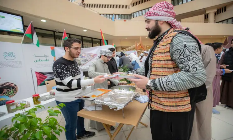 يستلم رجل مرتدياً غترة وشماغاً وعقالاً وبشتاً سعودياً تلذذ من المأكولات من بين منصات في مهرجان تراث الشعوب، حيت حيث الأعلام والموروثات الفلسطينية التراثية، بينما يحتفي طلاب القلم وزوار بحضارات غنيتهم في الخلفية.
