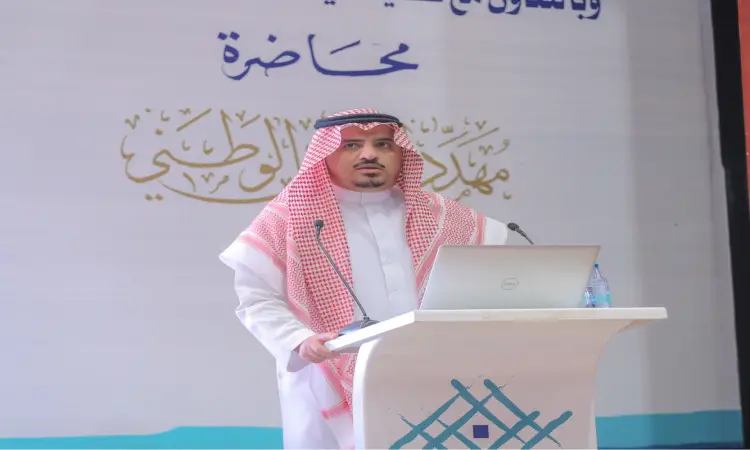 رجل يرتدي مرتديًا ثوبًا أبيض، وشماغًا سعوديًا وأبيض، مع عقال، ويرتدي أحمر بشتًا سعوديًا أنيقًا، خلف منصة كمبيوتر آلي محمول. غالباً تساعدها كلية نايف للأمن الوطني في القصيم. خذ وراءه لوحة تحمل الكتب باللغة العربية حول مهددات الأمن الوطني.