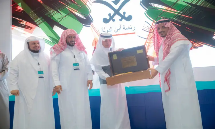 لأربعة رجال مرتدين الغترة والعقال والبشت السعودي على المسرح. هو أحد بتسليم درع محفوظ داخل المحافظة إلى رجل آخر. كوك خلفهم على شاشة شعار كلية نايف للأمن الوطني والسيفان المتقاطعان، وليس بعبارات باللغة العربية.