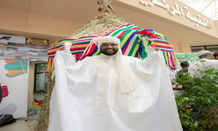 وقف رجل مبتسم مرتدياً غترة بيضاء وبشتاً سعودياً تقليدياً، ورافعاً رمزيه في مهرجان تراث الشعوب أمام مُجَسَّمٍ قشّي ملوّن خارج حرم جامعة القصيم، حيث حدت كتابات عربية أعلى المدخل.