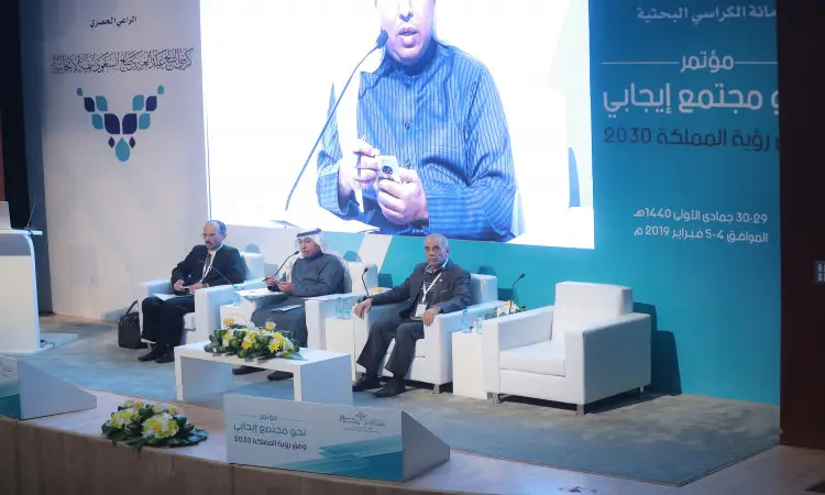 يجلس الرجال على المنصة خلال مؤتمر مجتمعي للترفيه، ويتناولون خلفهم لوحة كبيرة تحمل نصوصًا عربية وشعارًا لرؤية المملكة 2030. يتقدم أحد إلى المنبر وهو يرتدي البشت السعودي والغترة أو الشماغ مع العقال، بينما يجلس على كراسٍ أبيض في القاعة يزينها باقات الزهور على الطاولات؛ وذلك في إطار الفعاليات المقامة في القصيم.