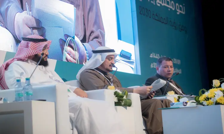 يجلس ثلاثة رجال على المنصة خلال مؤتمر رؤية 2030 في القصيم، ويرتدي اثنان منهم الغترة والعقال والبشت السعودي، بينما يرتدي الثالث بزّة رجالي. تتزين أمامهم برائحة النبيذ والزجاج. المؤقتة خلفهم على الشاشة الكبيرة تفاصيل الجلسات العلمية المكتوبة باللغة العربية.