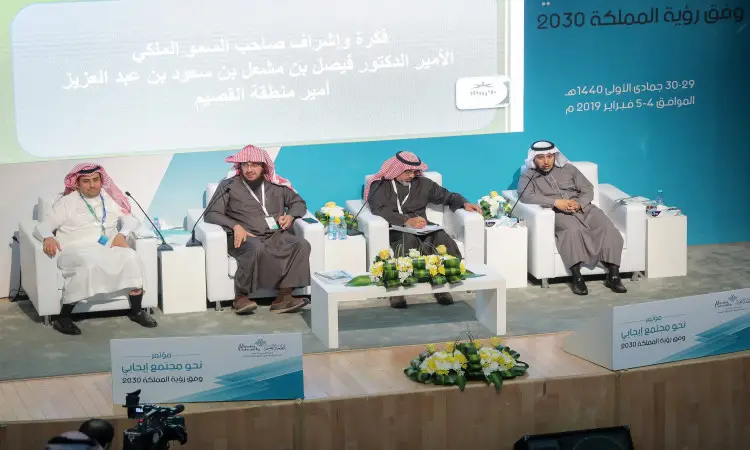 يجلس أربعة رجال ينتظرون البشتاء السعودي والغترة أو الشماغ والعقال خلال مؤتمر رؤية 2030 في جامعة القصيم، وتحيط بهم كراسي بيضاء وزهور جديدة. وتبرز خلفهم اللافتات التي تكتب عليها عبارات مثل "المجتمع الإيجابي" و"الجلسات العلمية"، إضافة إلى شعار رؤية 2030 العروض كدعوة للمؤتمر.