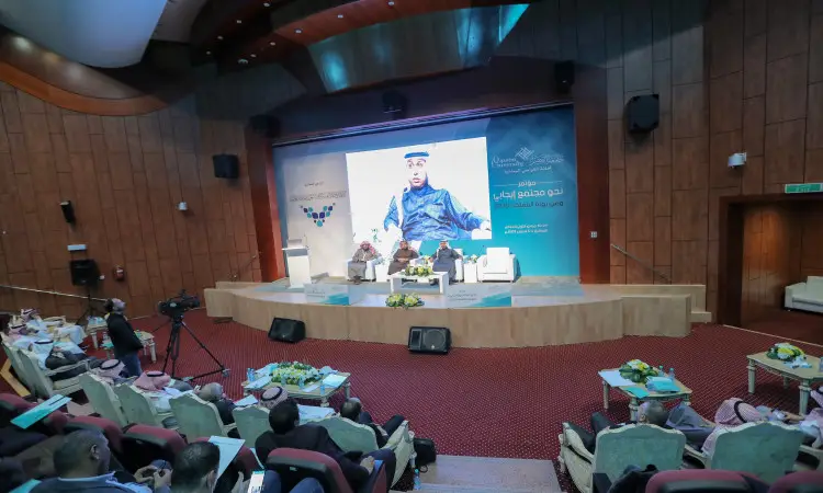 تنعقد حلقة نقاشية على المنصة في القاعة الكبرى، حيث يتم تحديد الجلسات العلمية للجمهور متابعاً باهتمامهم. لاحظ خلفين لوح الضغط المتوسط النصوصياً باللغة العربية وصورة لرجل مرتدياً الغترة السعودية والعقال والبشت الرسمي السعودي.