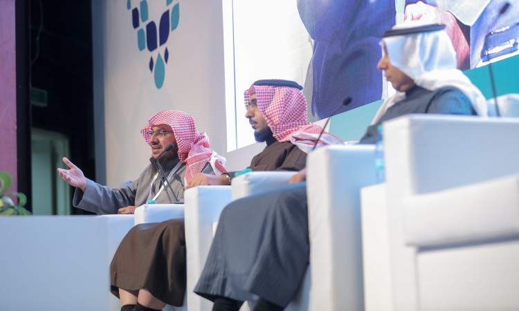يجلس ثلاثة رجال تحت الغترة أو الشماغ السعودي مع العقال والبشت السعودي على منصة المسرح فوق كراسٍ بيضاء، ويشاركون في جلسة حوارية ضمن المؤتمر الرسمي. يظهر أحد الأشخاص وهو يلوح بيده أثناء حديثه، بينما تم عرض شاشة كبيرة في المملكة العربية السعودية لرؤية 2030.